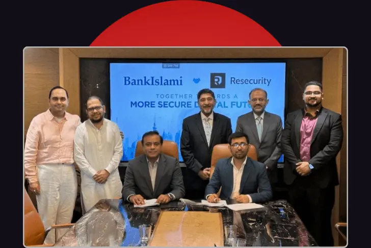 A formal group photograph of seven men gathered in a conference room to commemorate a corporate partnership. Two men are seated at a glossy conference table in the foreground, with five men standing behind them. In the background, a large digital screen displays the text 'BankIslami' and 'Resecurity' separated by a handshake icon, above the slogan 'TOGETHER TOWARDS A MORE SECURE DIGITAL FUTURE'. The photo is set against a dark background with a red semi-circle partially visible at the top edge