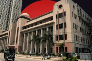 A composite image featuring the historical Cotton Exchange Building, a multi-story classical structure with tall columns and a small dome, set against a dark sky dominated by a massive solid red circle. A modern, textured white high-rise building is partially visible on the left. In the foreground, street-level activity includes a few pedestrians and green auto-rickshaws.