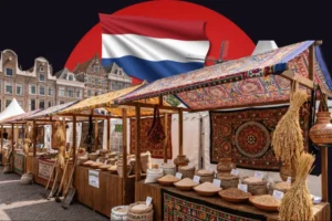 A composite image set against a dark background featuring a large red circle. At the top center, the flag of the Netherlands waves prominently. The main scene below shows an outdoor market with wooden stalls covered by ornately patterned, colorful fabric canopies. The stalls are richly stocked with various grains, seeds, and agricultural goods displayed in burlap sacks, shallow bowls, and clay pots, and are decorated with hanging dried wheat sheaves and traditional tapestries. In the background on the left, classic Dutch brick buildings with stepped gables are visible.