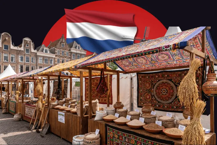A composite image set against a dark background featuring a large red circle. At the top center, the flag of the Netherlands waves prominently. The main scene below shows an outdoor market with wooden stalls covered by ornately patterned, colorful fabric canopies. The stalls are richly stocked with various grains, seeds, and agricultural goods displayed in burlap sacks, shallow bowls, and clay pots, and are decorated with hanging dried wheat sheaves and traditional tapestries. In the background on the left, classic Dutch brick buildings with stepped gables are visible.