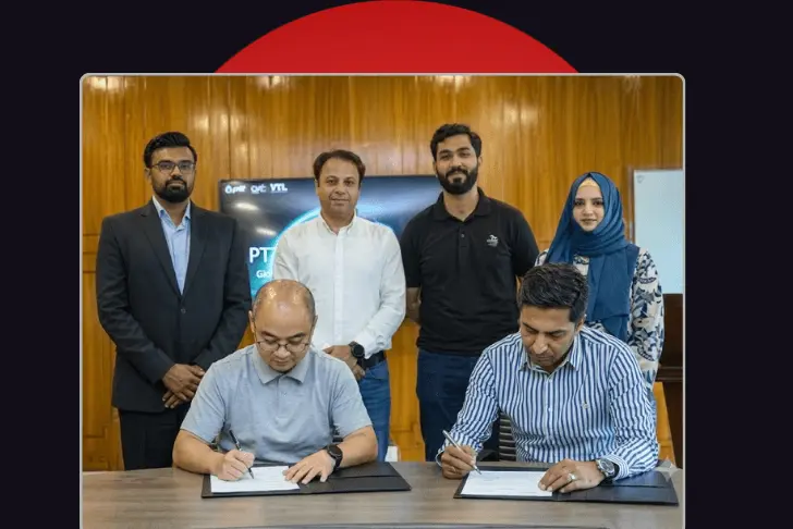 A group of six professionals gathered in a wood-paneled room for a formal signing event. In the foreground, two men are seated at a desk, simultaneously signing documents. Standing behind them are four colleagues—three men and one woman wearing a blue hijab—looking toward the camera. A digital screen displaying corporate logos is partially visible in the background. The photo is framed against a dark background with a red semi-circle at the top.