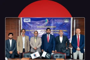 A formal group photograph showing six men in business suits standing in a row behind a conference table. Behind them hangs a blue banner that reads 'SIGNING CEREMONY' and displays the logos for DMT and PPL. On the table in the foreground, small flags for PPL and Pakistan sit next to document folders. The photograph is framed against a dark background with a large red semi-circle at the top.