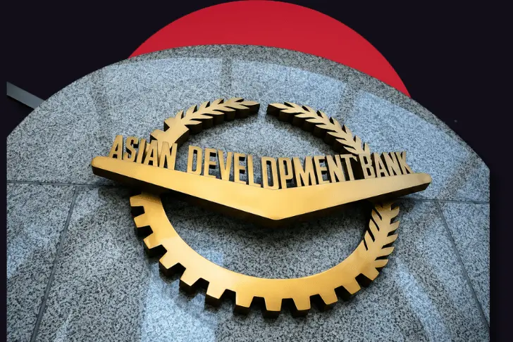 A close-up view of the Asian Development Bank logo, featuring three-dimensional gold lettering and a gold emblem combining a gear and laurel wreath, mounted on a curved grey stone surface. The top edge of the stone is set against a dark background with a partially visible, large red circle.