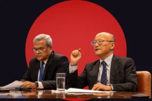 Two men in business suits seated at a glossy conference table. The man on the right, wearing glasses and a patterned tie, is looking forward and gesturing with his right hand as if making a point. The man on the left, also wearing glasses, is looking down to read from a document in his hands. The subjects are stylized against a dark background dominated by a massive, solid red circle positioned directly behind them.