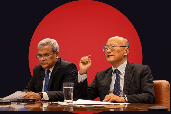 Two men in business suits seated at a glossy conference table. The man on the right, wearing glasses and a patterned tie, is looking forward and gesturing with his right hand as if making a point. The man on the left, also wearing glasses, is looking down to read from a document in his hands. The subjects are stylized against a dark background dominated by a massive, solid red circle positioned directly behind them.