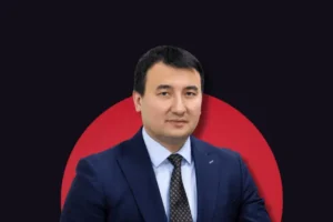 A portrait of a man wearing a dark blue suit, light blue shirt, and patterned tie, looking directly forward. He is set against a dark background with a large, solid red circle positioned directly behind him.