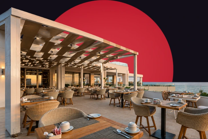 An outdoor restaurant patio overlooking the sea, featuring modern white pergolas, wooden tables, and woven wicker chairs set with tableware. The scene is set against a dark background with a massive, solid red circle rising behind the structure.