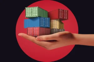 A stylized composite image showing a realistic human hand cradling a small stack of colorful miniature shipping containers (green, blue, red, yellow, and grey). The hand and containers are centered against a dark background featuring a large, solid red circle.