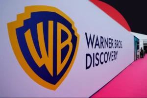 A large white wall displaying the blue-and-yellow Warner Bros. shield logo and the text 'WARNER BROS. DISCOVERY', positioned alongside a pink carpet walkway with a partial red graphic element in the upper right.