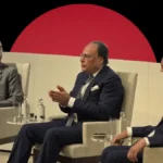 Three business executives seated on stage in panel chairs discussing economic policy during a formal conference, set against a dark background featuring a massive, solid red circle.