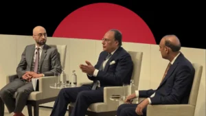 Three business executives seated on stage in panel chairs discussing economic policy during a formal conference, set against a dark background featuring a massive, solid red circle.