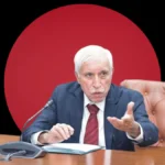 A professional portrait of Qaiser Ahmed Sheikh seated at a conference table in a dark blue suit and red tie, gesturing while speaking, set against a dark background featuring a massive solid red circle.