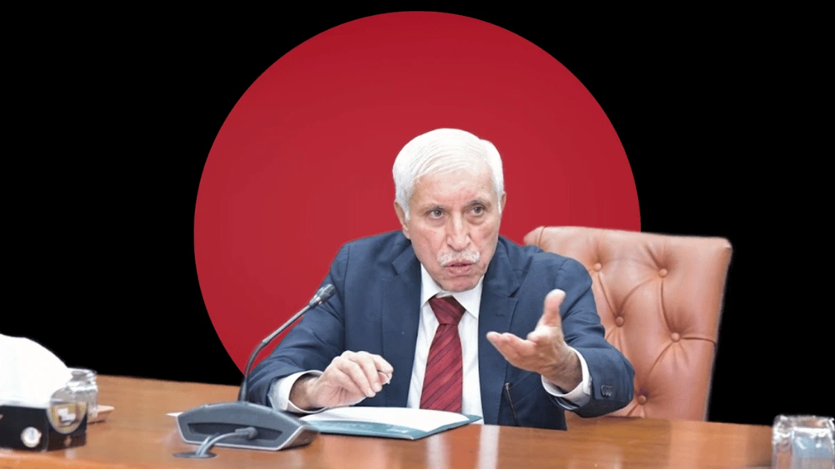 A professional portrait of Qaiser Ahmed Sheikh seated at a conference table in a dark blue suit and red tie, gesturing while speaking, set against a dark background featuring a massive solid red circle.